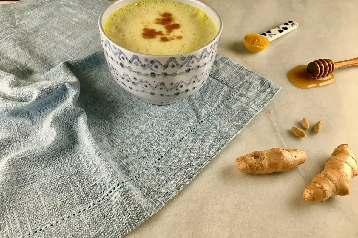 Bol de lait doré au curcuma, racines de gingembre et cuillère de miel sur un linge gris