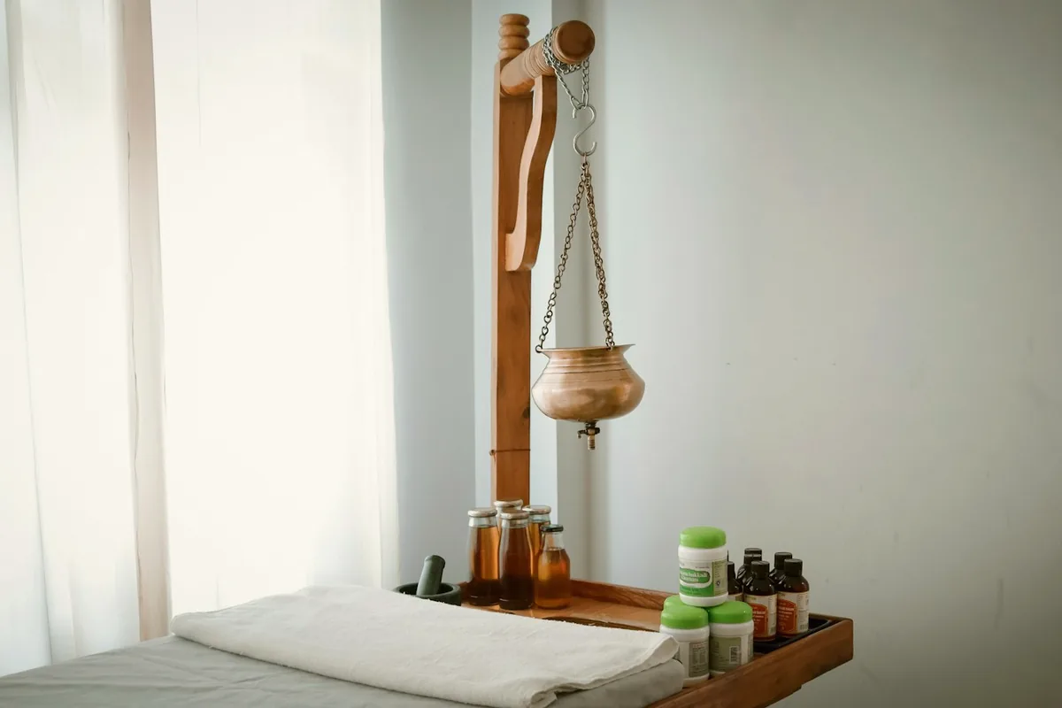 Salle de soin ayurvédique sobre : table recouverte de linge blanc, vase en cuivre suspendu pour le shirodhara, flacons d’huile